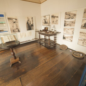 Fotografia de sala expositiva com vários quadros e fotografias históricas expostos nas paredes. Alguns itens estão expostos em mobiliários antigos. As paredes são brancas, em contraste com os pisos e móveis de madeira escura.