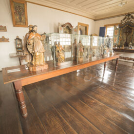 Fotografia de sala expositiva com paredes e teto claros e piso de madeira. Ao centro, há uma mesa de madeira com esculturas de santos expostas, algumas cobertas por vitrines de vidro. Ao fundo, vislumbram-se paredes brancas e um painel de madeira decorado, com imagens religiosas. A iluminação do ambiente é natural e suave.