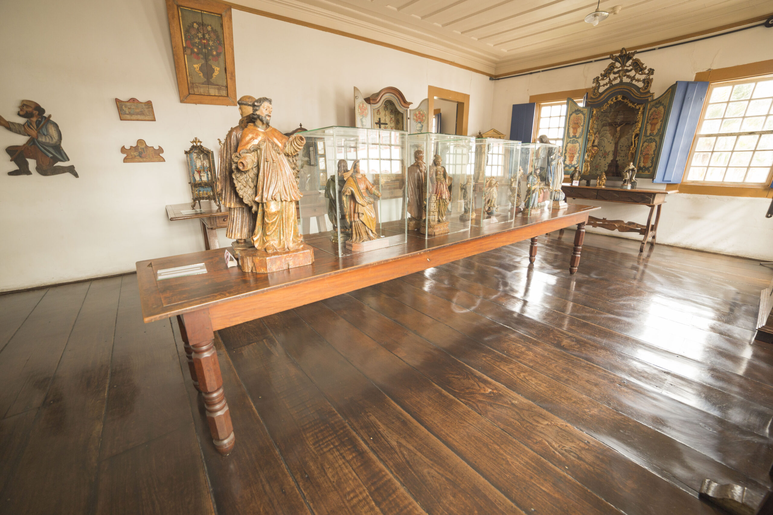 Fotografia de sala expositiva com paredes e teto claros e piso de madeira. Ao centro, há uma mesa de madeira com esculturas de santos expostas, algumas cobertas por vitrines de vidro. Ao fundo, vislumbram-se paredes brancas e um painel de madeira decorado, com imagens religiosas. A iluminação do ambiente é natural e suave.