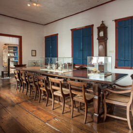 Fotografia de sala expositiva do museu, onde se localiza ao centro uma grande mesa de jantar, em madeira e com 12 cadeiras de madeira ornadas. Sob o tampo da mesa, há duas vitrines com objetos em destaque. A sala tem parede e teto claros e piso de madeira. Um relógio de parede fica entre as janelas fechadas. A luz do cômodo é central e difusa.