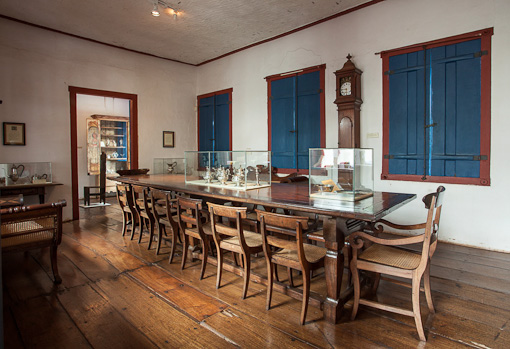 Fotografia de sala expositiva do museu, onde se localiza ao centro uma grande mesa de jantar, em madeira e com 12 cadeiras de madeira ornadas. Sob o tampo da mesa, há duas vitrines com objetos em destaque. A sala tem parede e teto claros e piso de madeira. Um relógio de parede fica entre as janelas fechadas. A luz do cômodo é central e difusa.