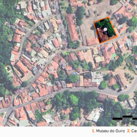 Vista de satélite da área do museu e seu entorno, mostrando a malha urbana com distribuição de casarios históricos e vegetações. Dois pontos estão em destaque, sendo o número 1, o Museu do Ouro, à direita da imagem, e o 2, localizado à esquerda, indicado como Casa Borba Gato. A imagem inclui a legenda com a identificação das edificações, no canto inferior direito, a escala gráfica e, na parte superior esquerda, uma seta indicando o ponto cardeal norte.