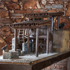 Fotografia da exposição, mostrando uma maquete detalhada de um moinho de água. A estrutura é feita de madeira, coberta por telhado de barro em duas alturas. No seu interior, existe uma roda d'água central. O ambiente onde está a maquete possui parede de pedras aparentes.