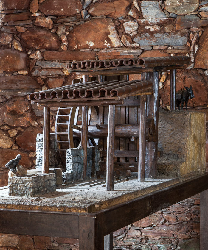 Fotografia da exposição, mostrando uma maquete detalhada de um moinho de água. A estrutura é feita de madeira, coberta por telhado de barro em duas alturas. No seu interior, existe uma roda d'água central. O ambiente onde está a maquete possui parede de pedras aparentes.