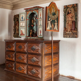 Fotografia de sala de exposição. A sala tem paredes e teto claros, com piso de madeira polida. Há uma cômoda de madeira exposta, com nove gavetas. Sobre a cômoda, está exposto um oratório religioso com as portas abertas, exibindo pequenas estátuas e figuras pintadas à mão. À direita do oratório, há uma pintura religiosa em pé sobre um suporte, retratando três figuras com vestes. Ao fundo, na parede, dois painéis entalhados em madeira, um de cada lado do oratório, com figuras religiosas em relevo.