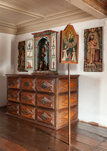 Fotografia de sala de exposição. A sala tem paredes e teto claros, com piso de madeira polida. Há uma cômoda de madeira exposta, com nove gavetas. Sobre a cômoda, está exposto um oratório religioso com as portas abertas, exibindo pequenas estátuas e figuras pintadas à mão. À direita do oratório, há uma pintura religiosa em pé sobre um suporte, retratando três figuras com vestes. Ao fundo, na parede, dois painéis entalhados em madeira, um de cada lado do oratório, com figuras religiosas em relevo.