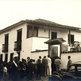 Fotografia antiga, que retrata uma cena histórica na fachada do Museu do Ouro. A edificação possui dois pavimentos e, em frente ao portão lateral, um grupo de pessoas está reunido, algumas entrando ou paradas próximas à entrada. Entre os presentes, há uma banda musical, com instrumentos visíveis como uma tuba, sugerindo um evento festivo ou cerimonial.