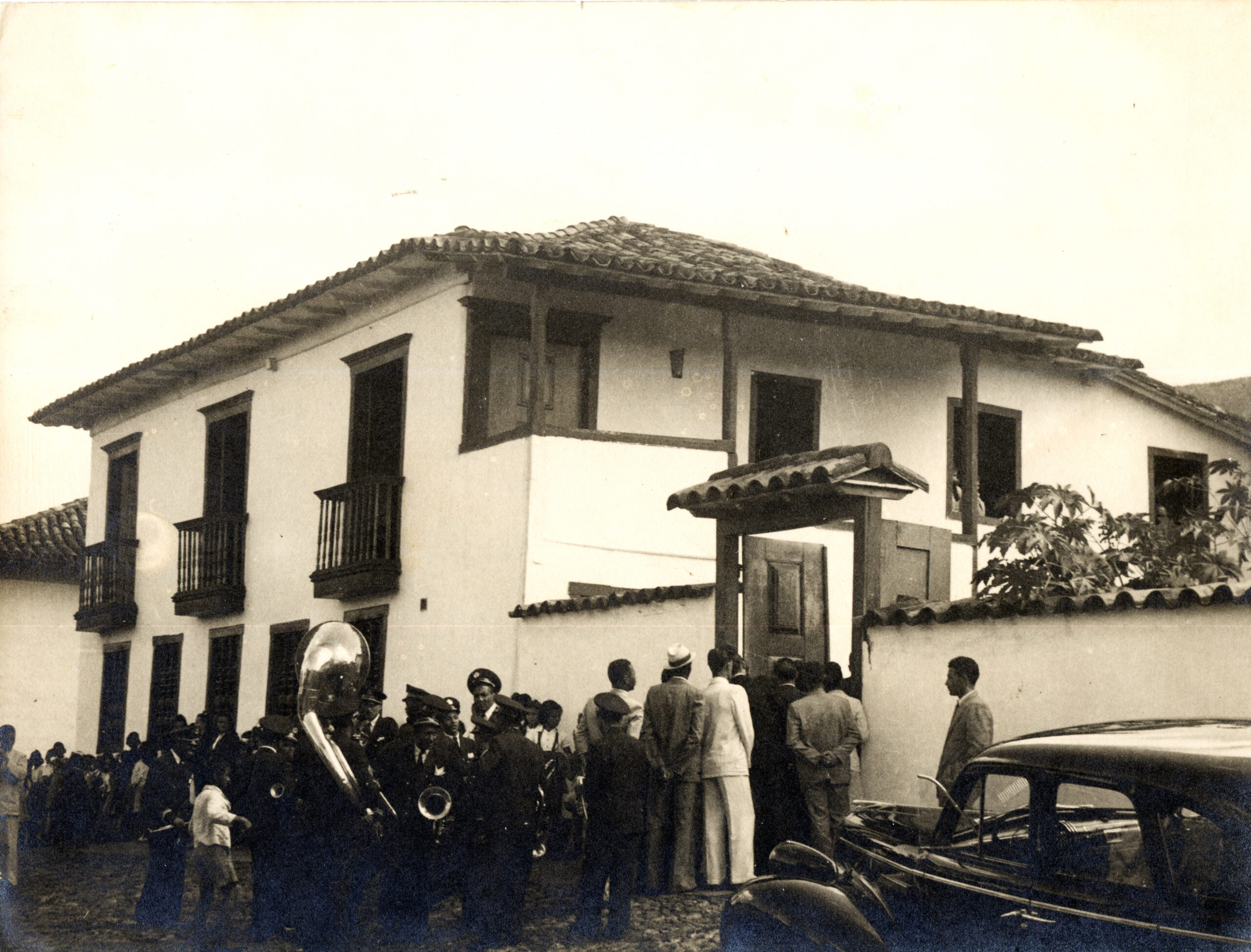 Fotografia antiga, que retrata uma cena histórica na fachada do Museu do Ouro. A edificação possui dois pavimentos e, em frente ao portão lateral, um grupo de pessoas está reunido, algumas entrando ou paradas próximas à entrada. Entre os presentes, há uma banda musical, com instrumentos visíveis como uma tuba, sugerindo um evento festivo ou cerimonial.