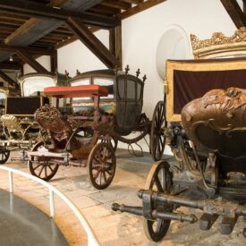 Fotografia da sala expositiva apresentando carruagens em uma área com piso rebaixado no ambiente e protegido por um guarda-corpo baixo. Cada carruagem possui elementos decorativos únicos e uma variedade de cores e acabamentos, incluindo madeira escura, dourados e materiais como tecido vermelho. Variam em tamanho e estilo.