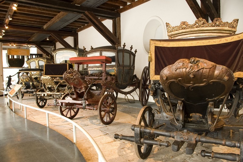 Fotografia da sala expositiva apresentando carruagens em uma área com piso rebaixado no ambiente e protegido por um guarda-corpo baixo. Cada carruagem possui elementos decorativos únicos e uma variedade de cores e acabamentos, incluindo madeira escura, dourados e materiais como tecido vermelho. Variam em tamanho e estilo.