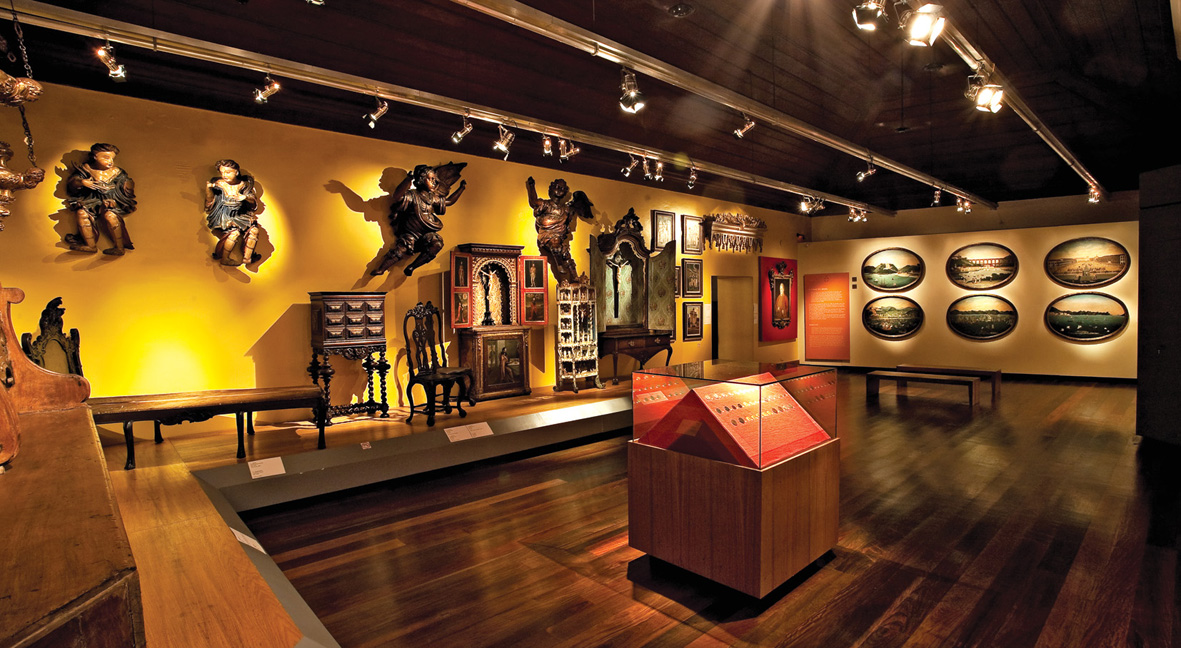 Fotografia de sala de exposição, com paredes pintadas de amarelo, teto escuro com trilhos de iluminação e piso de madeira lustrada. Ao centro, uma vitrine baixa destaca um objeto através de iluminação direcionada. Nas paredes, objetos pendurados formam uma composição de imagens sacras. Um tablado ladeia as paredes e sobre ele há mobiliários de épocas diferentes.