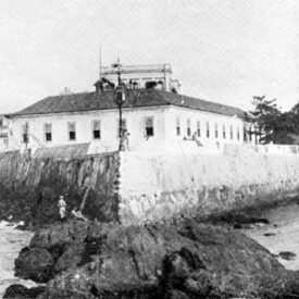 Imagem histórica em preto e branco. Mostra uma construção do forte de pedra branca. No primeiro plano, o mar, e no segundo, o platô de pedras no qual o edifício foi construído.
