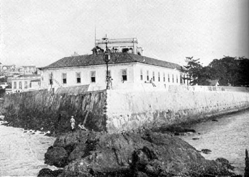 Imagem histórica em preto e branco. Mostra uma construção do forte de pedra branca. No primeiro plano, o mar, e no segundo, o platô de pedras no qual o edifício foi construído.