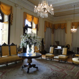 Fotografia de área expositiva, que consiste em uma sala de estar com pé-direito alto, teto ornamentado e dois lustres de cristal suspensos. A iluminação natural entra por grandes janelas com cortinas amarelas drapeadas. No centro do espaço, destaca-se um conjunto de móveis antigos, formado por um sofá longo e quatro cadeiras estofadas em amarelo, organizados ao redor de uma mesa redonda com um arranjo de flores brancas. O piso é revestido por um amplo tapete de desenhos florais.