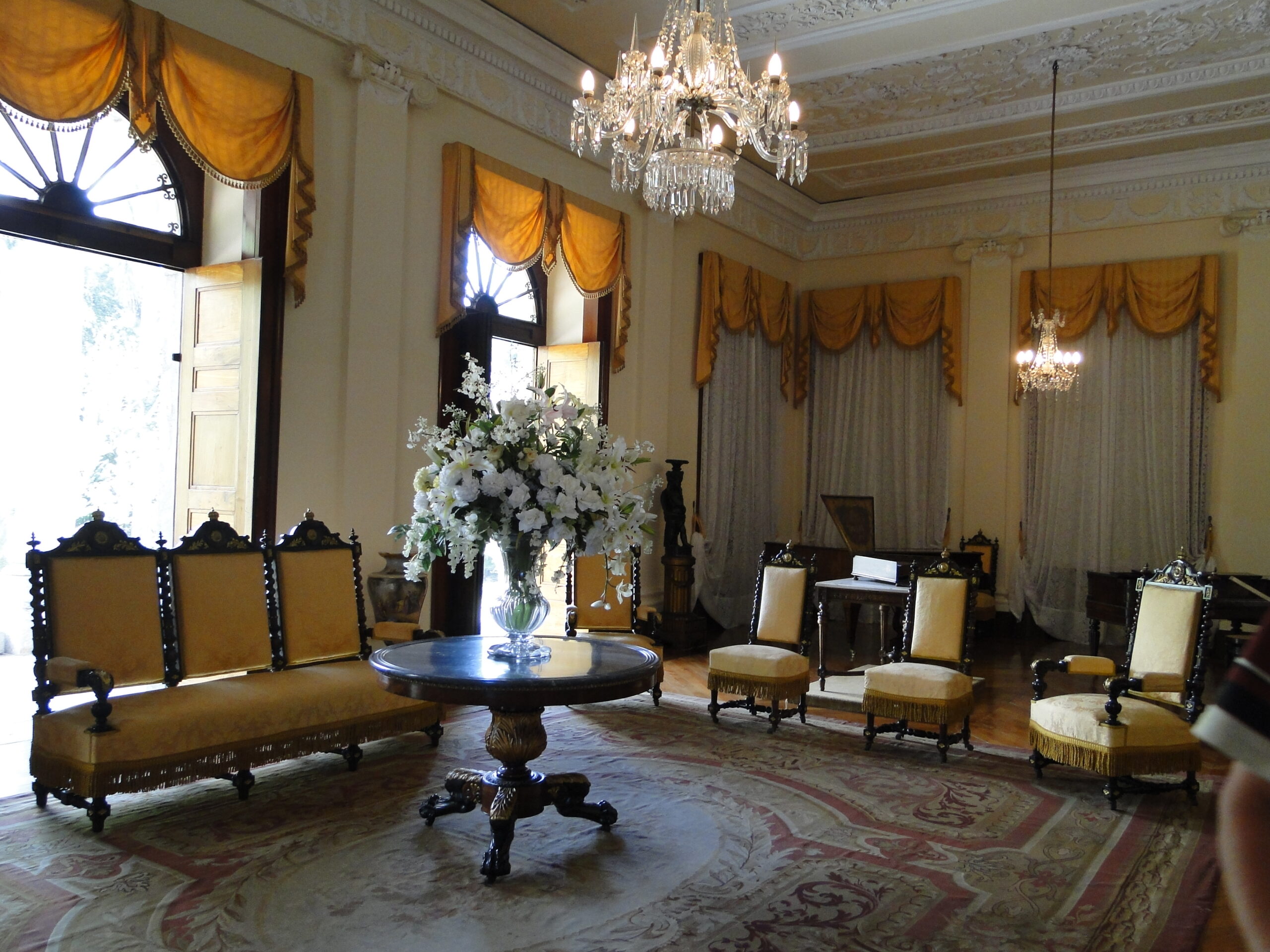 Fotografia de área expositiva, que consiste em uma sala de estar com pé-direito alto, teto ornamentado e dois lustres de cristal suspensos. A iluminação natural entra por grandes janelas com cortinas amarelas drapeadas. No centro do espaço, destaca-se um conjunto de móveis antigos, formado por um sofá longo e quatro cadeiras estofadas em amarelo, organizados ao redor de uma mesa redonda com um arranjo de flores brancas. O piso é revestido por um amplo tapete de desenhos florais.