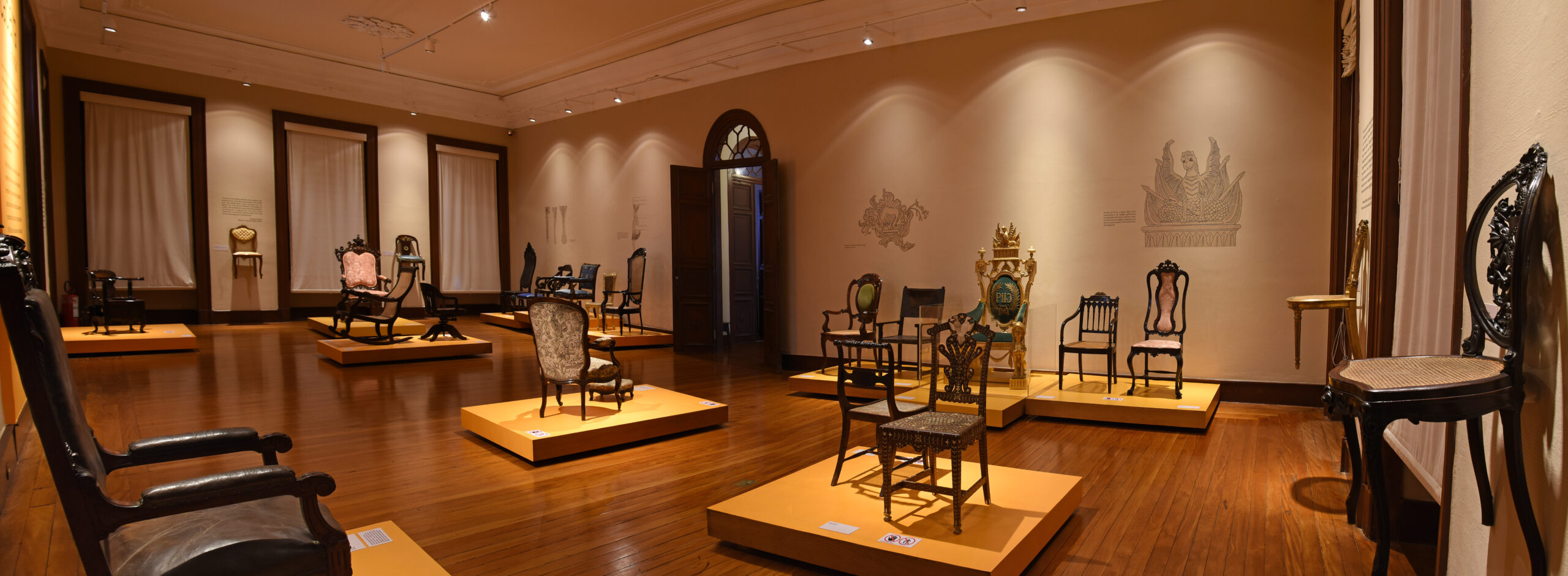 Fotografia da sala de exposição com piso de madeira e paredes claras, apresentando diversas cadeiras dispostas sobre bases retangulares. Cada cadeira possui uma placa descritiva à frente. Ao fundo, janelas com cortinas brancas filtram a luz natural, enquanto luminárias embutidas no teto complementam a iluminação. No centro da parede posterior, uma porta com arco dá acesso a outra sala do museu.