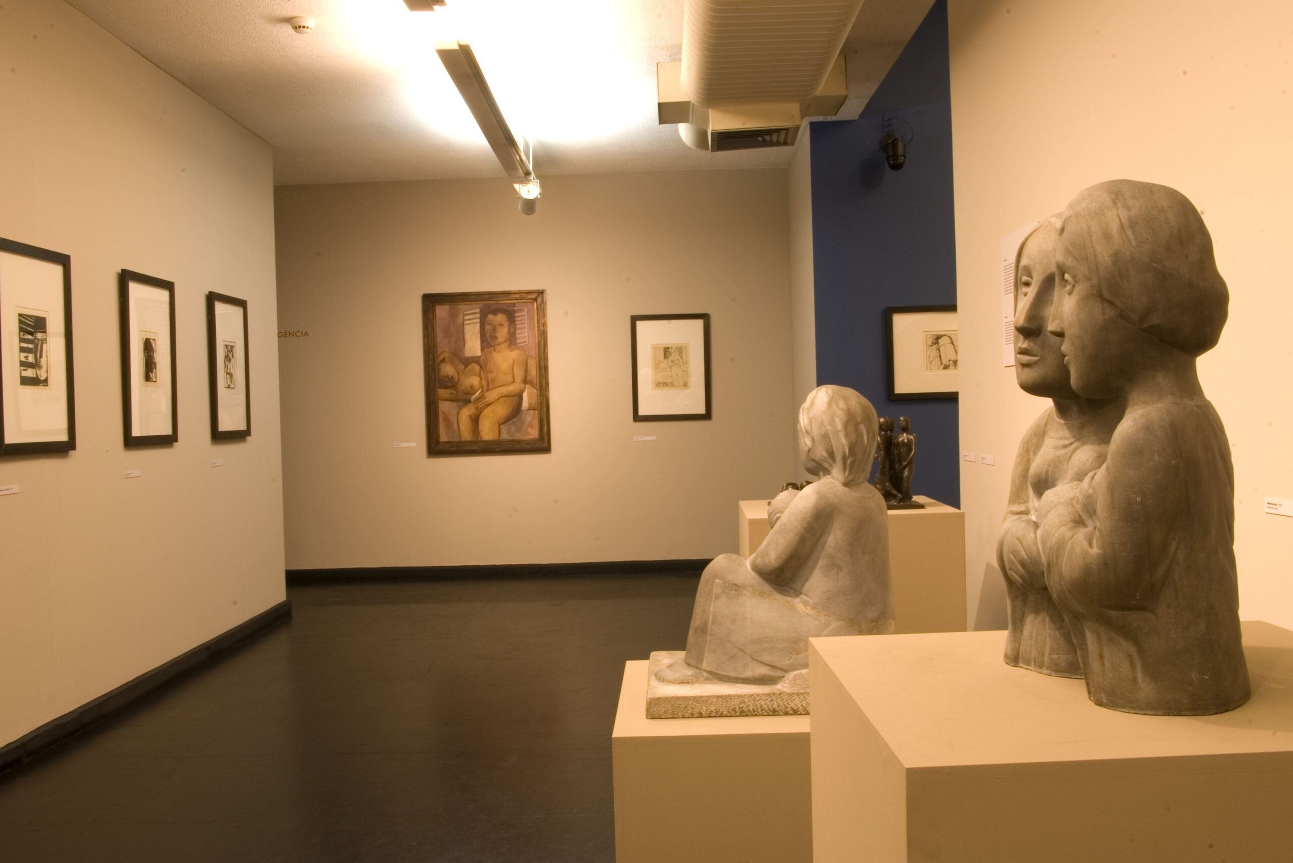Fotografia do interior do museu. Sala expositiva de parede clara e piso de madeira, com quadros alinhados de vários tamanhos distribuídos nas paredes. Ao fundo, no centro, há um quadro maior com a pintura de uma pessoa sentada. À direita, outro quadro menor, também emoldurado. Em primeiro plano, à direita, duas esculturas de figuras humanas sentadas, posicionadas sobre pedestais quadrados.