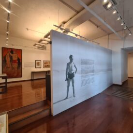 Fotografia de sala de exposição. A sala é ampla, dividida em dois ambientes com desníveis de piso. As paredes e o teto são claros, com trilhos distribuídos ao longo do espaço com luminárias destacando as obras. Um painel central expõe uma grande fotografia em preto e branco de um homem de pé, vestindo apenas calções. Ao lado da fotografia: um texto explicativo da imagem principal.