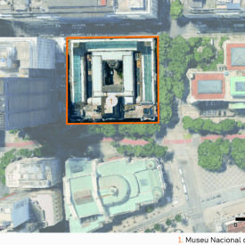 Vista aérea de um quarteirão urbano, com o museu ao centro, no alto da imagem, destacado por um retângulo laranja. Ao redor, há outros edifícios, ruas e áreas verdes visíveis. A imagem inclui a legenda com a identificação das edificações, no canto inferior direito, a escala gráfica e, na parte superior esquerda, uma seta indicando o ponto cardeal norte.
