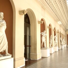 Sala de exposição do museu, com dimensões amplas de comprimento e altura. Possui paredes claras com acabamentos em arcos na tonalidade marrom. Os arcos e colunas são dispostos de maneira simétrica na parede, destacando estátuas de mármore que retratam figuras humanas em diversas posições. O teto alto possui um acabamento abobadado, com uma claraboia que facilita a entrada de luz natural. O piso claro favorece a iluminação e a impressão de espaço amplo.