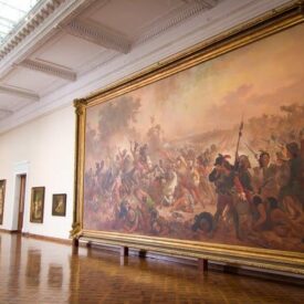Fotografia de sala expositiva. A sala tem formato retangular e amplo, com pé-direito alto, paredes claras, teto branco, piso de madeira e iluminação zenital proveniente da cobertura de vidro. No centro da sala, há bancos em frente a um grande quadro pendurado em uma das paredes. Com moldura dourada e pintura a óleo, a obra retrata uma batalha histórica.