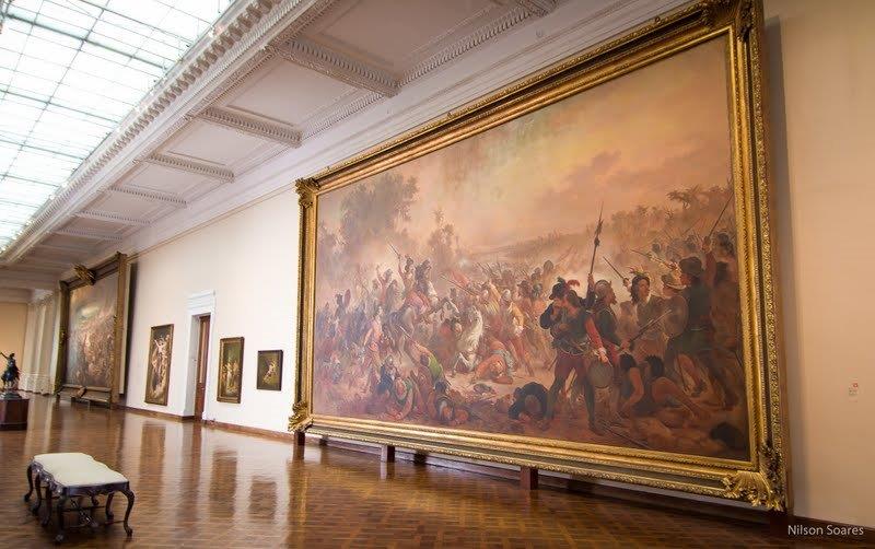 Fotografia de sala expositiva. A sala tem formato retangular e amplo, com pé-direito alto, paredes claras, teto branco, piso de madeira e iluminação zenital proveniente da cobertura de vidro. No centro da sala, há bancos em frente a um grande quadro pendurado em uma das paredes. Com moldura dourada e pintura a óleo, a obra retrata uma batalha histórica.