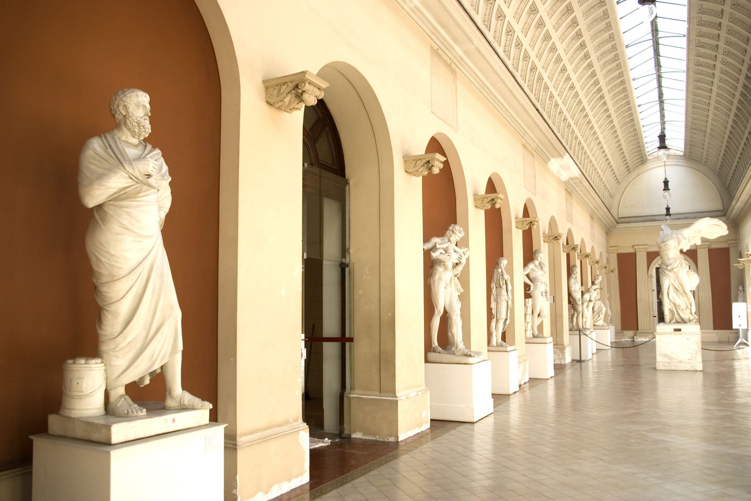 Sala de exposição do museu, com dimensões amplas de comprimento e altura. Possui paredes claras com acabamentos em arcos na tonalidade marrom. Os arcos e colunas são dispostos de maneira simétrica na parede, destacando estátuas de mármore que retratam figuras humanas em diversas posições. O teto alto possui um acabamento abobadado, com uma claraboia que facilita a entrada de luz natural. O piso claro favorece a iluminação e a impressão de espaço amplo.