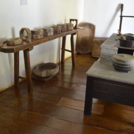 Sala de exposição, com paredes brancas e piso de madeira. Na parede, um aparador com diversos utensílios e ferramentas de preparo de alimentos. No centro, uma mesa baixa contendo outros utensílios de ferro e pedra.