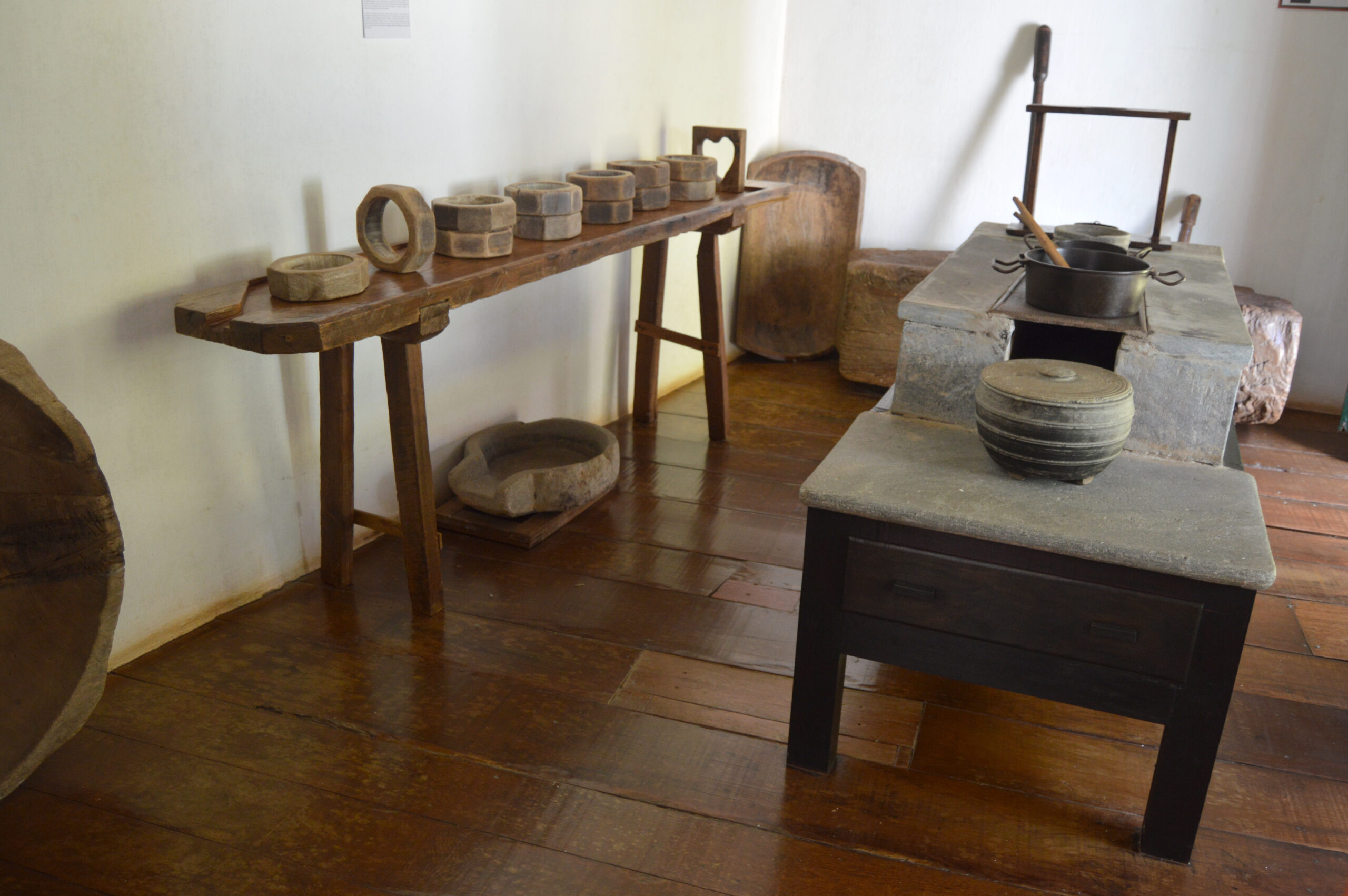 Sala de exposição, com paredes brancas e piso de madeira. Na parede, um aparador com diversos utensílios e ferramentas de preparo de alimentos. No centro, uma mesa baixa contendo outros utensílios de ferro e pedra.