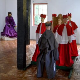 Sala de exposições. Exposição com imagens de roca, trata-se de imagens sacras que se destinam a ser levadas em procissão e que são vestidas com trajes de tecido. Estão representados Jesus Cristo, com vestes roxas; com vestes vermelhas e brancas, cardeais circundam um bispo da Igreja Católica e, vestido com um hábito religioso marrom, está representado São Francisco de Assis.