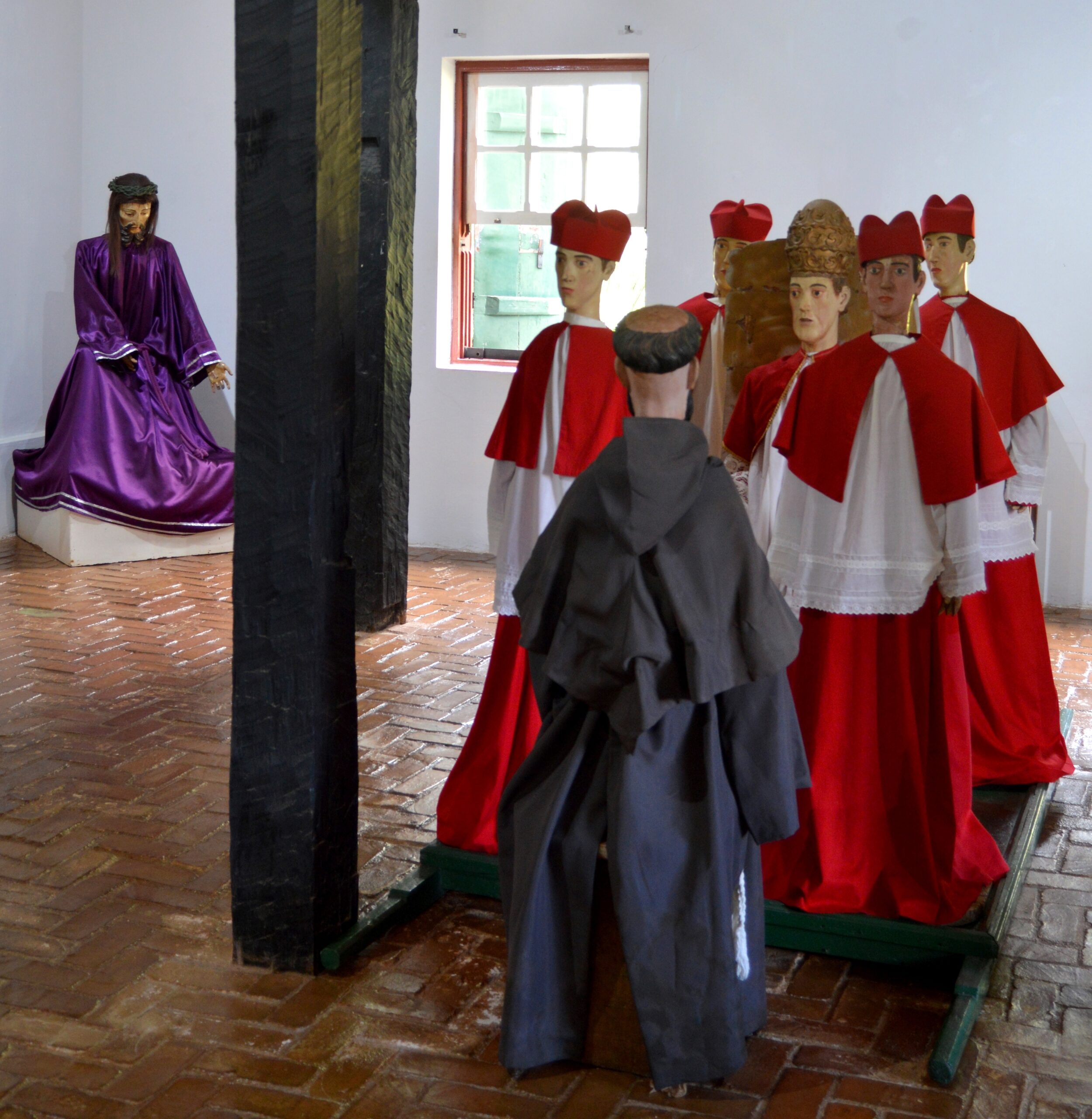 Sala de exposições. Exposição com imagens de roca, trata-se de imagens sacras que se destinam a ser levadas em procissão e que são vestidas com trajes de tecido. Estão representados Jesus Cristo, com vestes roxas; com vestes vermelhas e brancas, cardeais circundam um bispo da Igreja Católica e, vestido com um hábito religioso marrom, está representado São Francisco de Assis.