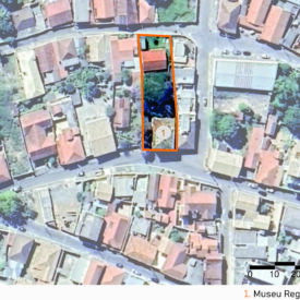 Vista aérea da área urbana com várias construções e poucas áreas verdes. As ruas acompanham o desenho natural do terreno. No centro, há um contorno laranja destacando o terreno do museu. A imagem inclui a legenda com a identificação das edificações, no canto inferior direito, a escala gráfica e, na parte superior esquerda, uma seta indicando o ponto cardeal norte.