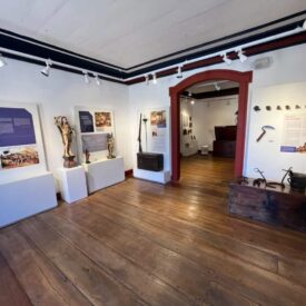 Fotografia da sala expositiva, com paredes brancas e piso de madeira. A iluminação uniforme e bem distribuída destaca as peças sem causar sombras fortes, enfatizando os detalhes históricos expostos em vitrines e painéis. Os objetos e peças históricas incluem esculturas, armas e ferramentas antigas, bem como painéis informativos.