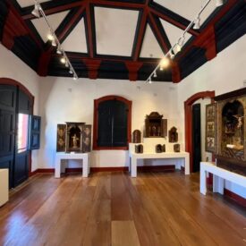 Fotografia de sala de exposição, de teto alto com detalhes do forro pintado em azul e vermelho, criando um contraste marcante. A sala tem janelas e portas com molduras, destacando os planos e o piso de madeira polida. Três mesas brancas ao longo das paredes exibem diferentes oratórios. À esquerda, há uma escultura de madeira sobre um pedestal branco. À direita, há um oratório decorado com pinturas religiosas.