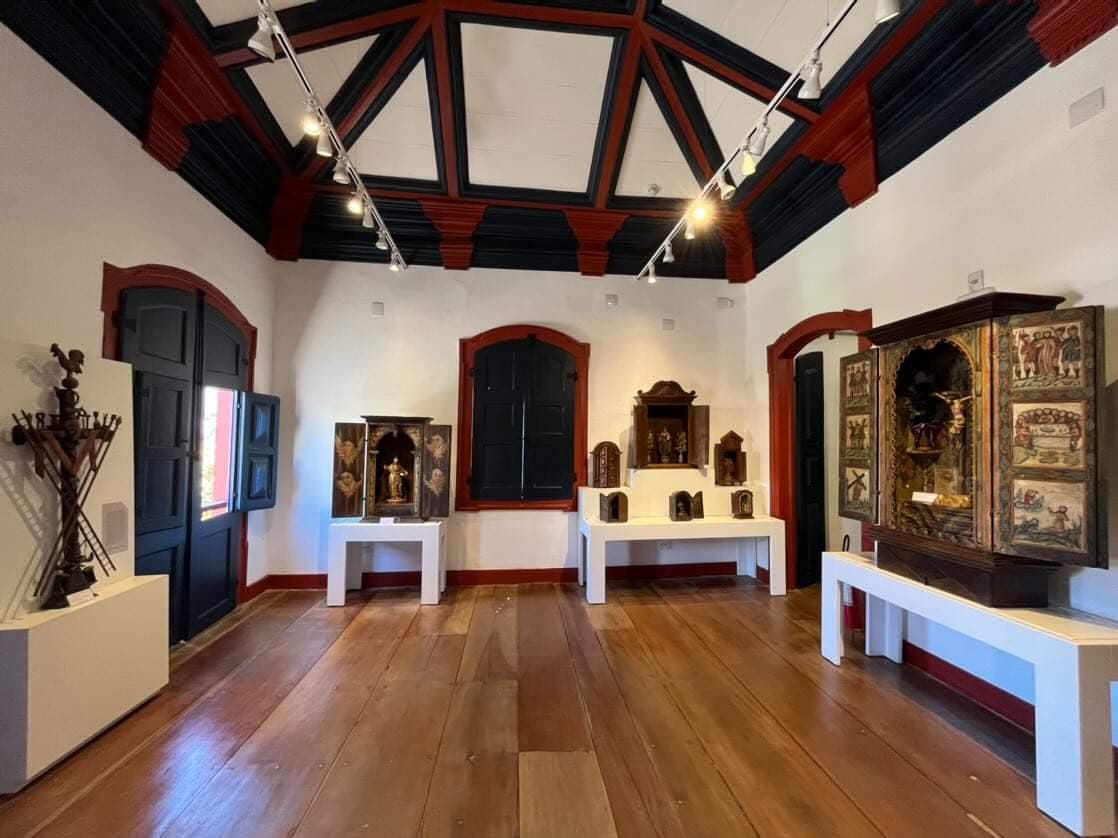 Fotografia de sala de exposição, de teto alto com detalhes do forro pintado em azul e vermelho, criando um contraste marcante. A sala tem janelas e portas com molduras, destacando os planos e o piso de madeira polida. Três mesas brancas ao longo das paredes exibem diferentes oratórios. À esquerda, há uma escultura de madeira sobre um pedestal branco. À direita, há um oratório decorado com pinturas religiosas.