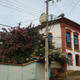 Fotografia antiga do Museu. A composição apresenta uma perspectiva de rua com declive, com o museu como elemento central. A casa se estende do primeiro plano ao fundo, com paredes brancas e detalhes em vermelho-tijolo, com janelas azuis e uma abundante vegetação de flores roxas em volta. O tempo está parcialmente ensolarado, com luz difusa e pouca sombra aparente.