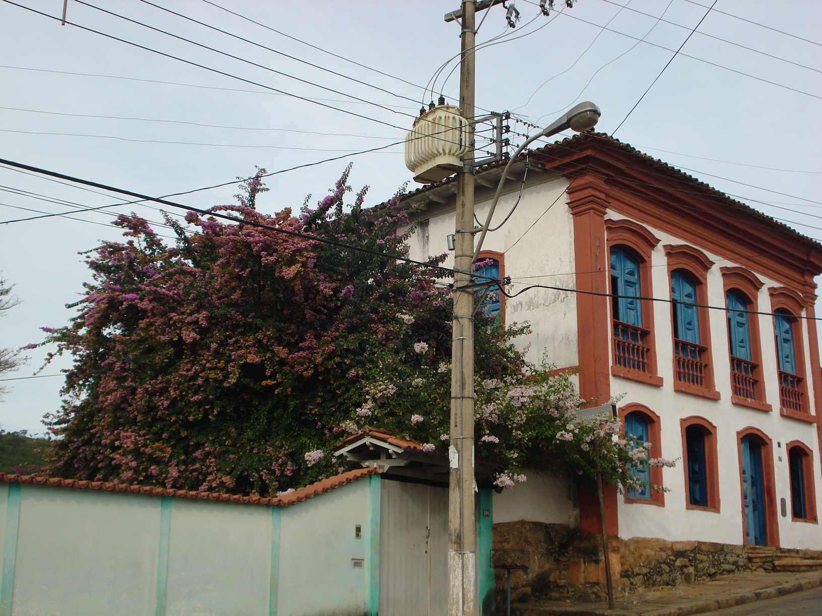 Fotografia antiga do Museu. A composição apresenta uma perspectiva de rua com declive, com o museu como elemento central. A casa se estende do primeiro plano ao fundo, com paredes brancas e detalhes em vermelho-tijolo, com janelas azuis e uma abundante vegetação de flores roxas em volta. O tempo está parcialmente ensolarado, com luz difusa e pouca sombra aparente.
