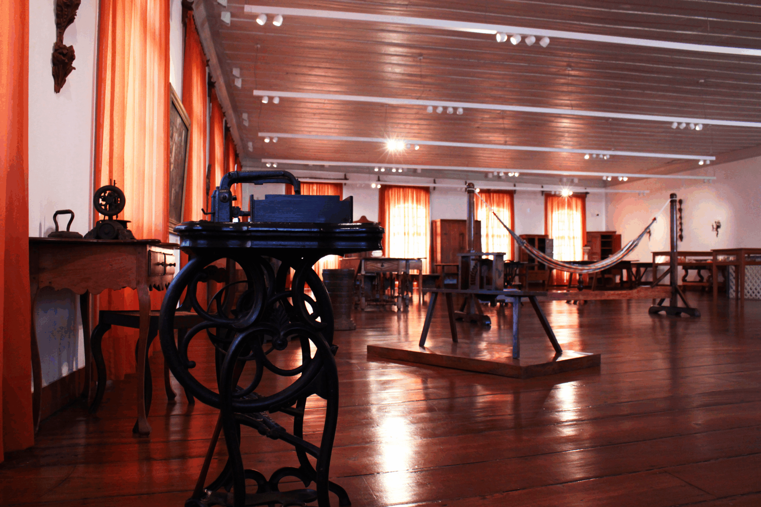 Fotografia da sala expositiva. Sala com piso de madeira polida, com contrastes suaves entre os tons claros das paredes e o tom alaranjado das cortinas. O espaço é amplo e há diversas peças de mobiliário, bem como instrumentos e utensílios, dispostos em diferentes partes da sala. A iluminação é natural, proveniente de janelas.