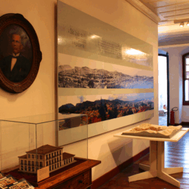 Sala expositiva com paredes e teto claros e piso de madeira polido. No primeiro plano, a parede destaca um painel com paisagem e, à esquerda da parede, um quadro oval com um retrato de uma pessoa. Há uma vitrine de vidro com a maquete do edifício em miniatura e um suporte com um livro. No fundo, em segundo plano, as janelas em arco batido que dão para a rua estão abertas e refletem a luz natural.