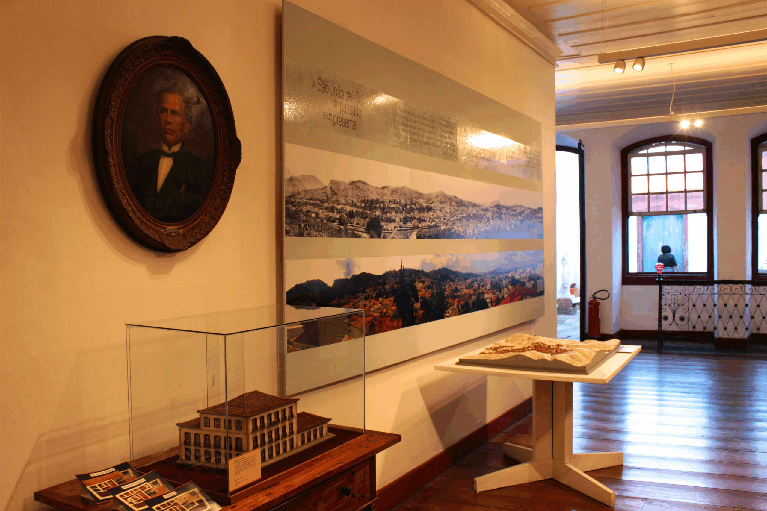 Sala expositiva com paredes e teto claros e piso de madeira polido. No primeiro plano, a parede destaca um painel com paisagem e, à esquerda da parede, um quadro oval com um retrato de uma pessoa. Há uma vitrine de vidro com a maquete do edifício em miniatura e um suporte com um livro. No fundo, em segundo plano, as janelas em arco batido que dão para a rua estão abertas e refletem a luz natural.