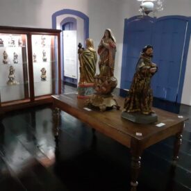 Fotografia de um espaço expositivo no interior do Museu. Ao centro, há uma mesa de madeira com esculturas sacras em destaque. À esquerda, uma vitrine exibe outras peças do acervo, também de temática religiosa. O ambiente possui piso de madeira escura, paredes claras e esquadrias em azul, incluindo janelas com folhas internas fechadas e arco superior.