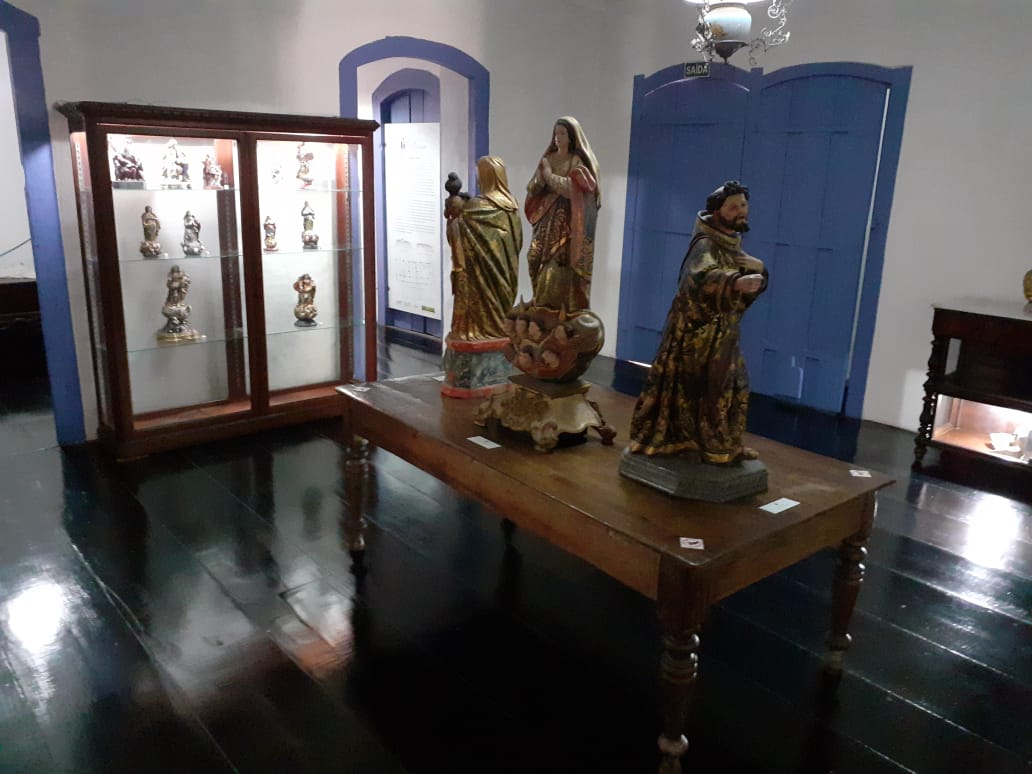 Fotografia de um espaço expositivo no interior do Museu. Ao centro, há uma mesa de madeira com esculturas sacras em destaque. À esquerda, uma vitrine exibe outras peças do acervo, também de temática religiosa. O ambiente possui piso de madeira escura, paredes claras e esquadrias em azul, incluindo janelas com folhas internas fechadas e arco superior.