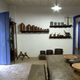Fotografia de um espaço expositivo do museu. O ambiente tem paredes brancas, piso de tijolos e uma mesa rústica de madeira com bancos e tigelas de cerâmica. À esquerda, há uma porta azul aberta e uma cristaleira com pratos. Na parede do fundo, prateleiras expõem objetos antigos como potes de cobre, xícaras e moedores. Mais ao fundo, está um corredor com portas de verga azul.