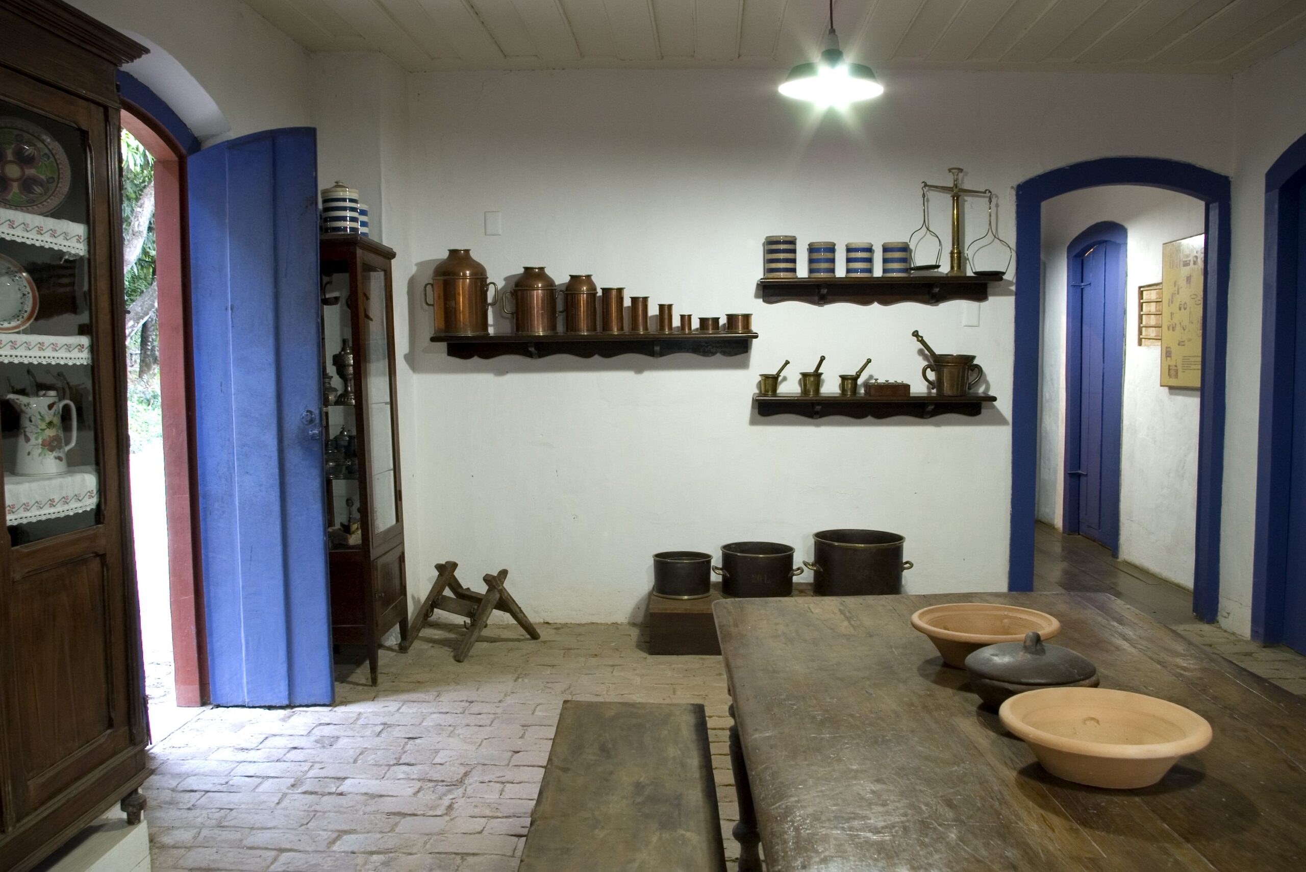 Fotografia de um espaço expositivo do museu. O ambiente tem paredes brancas, piso de tijolos e uma mesa rústica de madeira com bancos e tigelas de cerâmica. À esquerda, há uma porta azul aberta e uma cristaleira com pratos. Na parede do fundo, prateleiras expõem objetos antigos como potes de cobre, xícaras e moedores. Mais ao fundo, está um corredor com portas de verga azul.