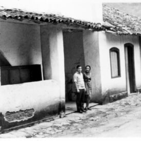 Fotografia histórica em preto e branco do Museu. A edificação possui fachada de alvenaria e cobertura de telhas cerâmicas aparentes. A casa possui uma sequência de aberturas retangulares que alternam entre portas e janelas. À esquerda da imagem, há um pequeno alpendre onde duas pessoas estão de pé, próximas uma da outra, olhando em direção à câmera. O piso em frente à casa é irregular.