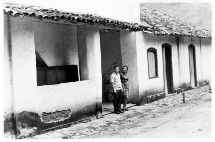 Fotografia histórica em preto e branco do Museu. A edificação possui fachada de alvenaria e cobertura de telhas cerâmicas aparentes. A casa possui uma sequência de aberturas retangulares que alternam entre portas e janelas. À esquerda da imagem, há um pequeno alpendre onde duas pessoas estão de pé, próximas uma da outra, olhando em direção à câmera. O piso em frente à casa é irregular.
