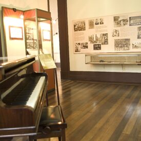 Fotografia da área expositiva. Há um piano de madeira polida em primeiro plano e, ao seu lado, uma vitrine expositiva. A parede à direita do piano está coberta por uma série de imagens em molduras pretas e brancas expostas em um fundo bege claro.