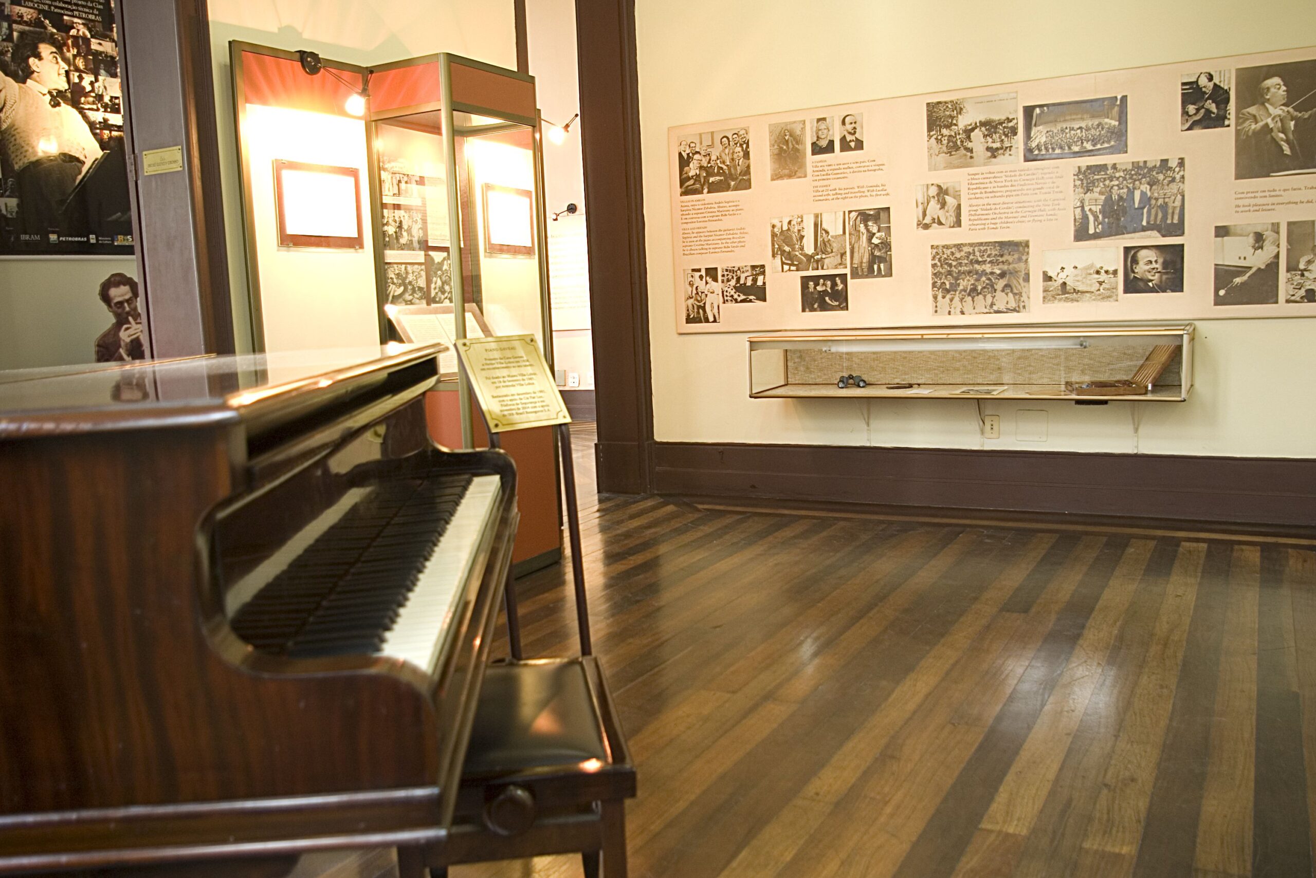 Fotografia da área expositiva. Há um piano de madeira polida em primeiro plano e, ao seu lado, uma vitrine expositiva. A parede à direita do piano está coberta por uma série de imagens em molduras pretas e brancas expostas em um fundo bege claro.
