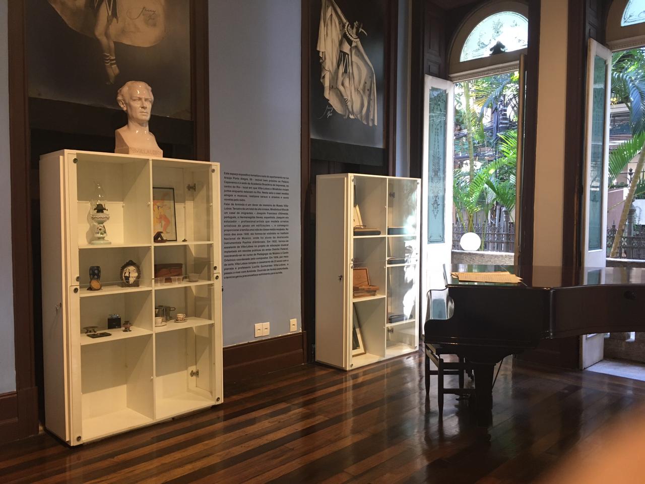 Fotografia da área expositiva. A composição da imagem apresenta uma sala com um piano preto posicionado no centro da cena, próximo à parede direita. Estantes brancas com objetos expostos ocupam a parede esquerda. A parede atrás da estante é de cor azul claro e uma moldura de madeira escura emoldura as paredes e o piso. No canto superior esquerdo, um busto de um homem. A luz natural entra pela janela dupla, localizada à direita do piano.