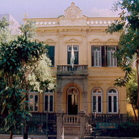 Fotografia da fachada do Museu. O edifício apresenta fachada em tons amarelados claros, com janelas e detalhes em moldura, criando uma composição simétrica. Há uma série de janelas, no térreo com bandeiras em arco pleno e no primeiro pavimento acabamento reto. Ao centro, acima da porta principal, um balcão ornado se sobressai do plano da fachada. Ao redor do prédio, vários arbustos e árvores verdes criam um contraste visual com a cor do edifício.